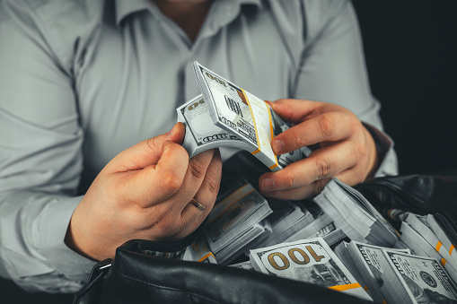 On a black background, a man is counting a wad of dollars over a bag of money. A man in a business shirt flips through dollar bills in his hand against a black background while sitting at a table.
