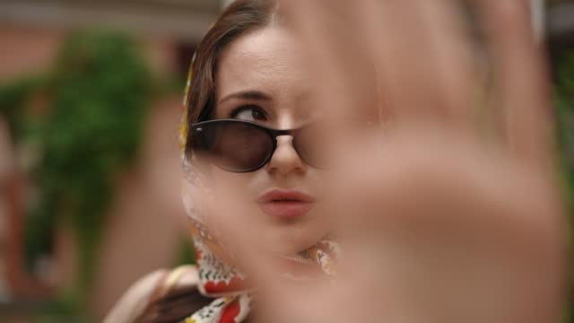 Slow motion. In the foreground, out of focus, is a woman's palm. In the background, a woman in a headscarf looks at the camera over her sunglasses while outdoors on a summer day.