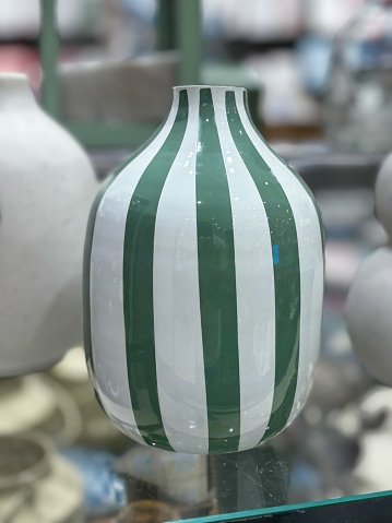 A green and white striped vase sits on a table. The stripes on the vase are bold and stand out against the white background. The vase is a statement piece that adds a pop of color to any room