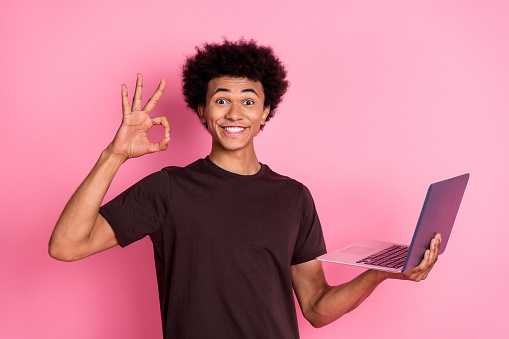 Photo of young shopaholic cheerful guy showing okey symbol holding netbook enjoy online shopping isolated on pink color background.