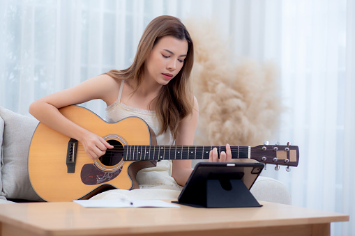 Young asian woman sitting on sofa looking tablet for practice music with guitar in living room at home, woman watching lesson online about music and learning guitar, lifestyle concept.