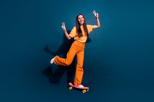 Full body size photo of smiling mature age hispanic woman has hobby riding skateboard makes v sign isolated on dark blue color background.