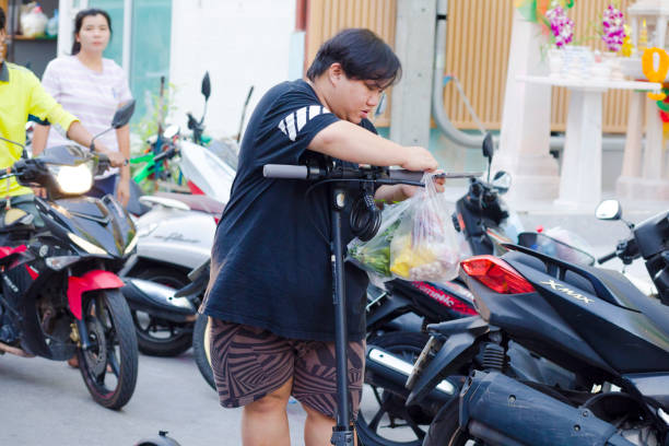Thai person with E-scooter and bag with vegetables  captured in street of residential district in Bangkok Chatuchak. In street arte market stalls of local market. Person has bags with bought food and vegetables and gettting onto scooter. Person is hanging bags at streering handles. In background are parked motorcycles and a woman