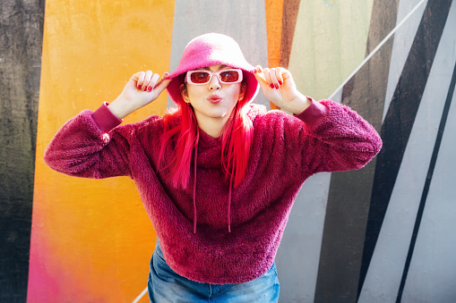 Hipster young woman with pink hair and sunglasses in magenta fluffy sweatshirt and bucket hat posing on the wall background. Urban street fashion. Mono color look. Gen Z, millennials self expression