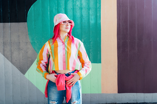 Young woman with pink hair and sunglasses in Bucket hat and multicolor strippled shirt posing on graffiti wall background. Street artist with mural, hipster, blogger. Urban street fashion.