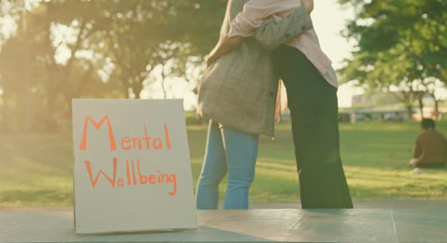 Two young Asian women sharing a supportive embrace in a public park during a mental health awareness event, highlighting compassion and care. Outdoor mental wellbeing community support.