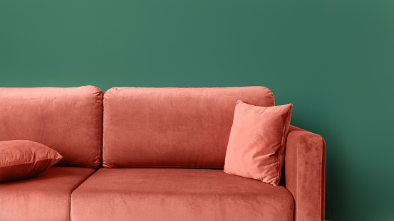 Close up of coral comfortable couch with cushions against green wall with copy space. Modern elegant furniture in living room at home. Clean, soft upholstery of sofa, web banner view