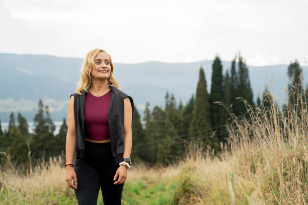 Young woman between 25-30 with short blonde hair and comfortable sports clothing walks through a field surrounded by wheat ears and with a forest in the background full of very tall trees and pines, she stops on the road smiling looking at the sky happy for a day of sports taking care of your health and enjoying the fresh air