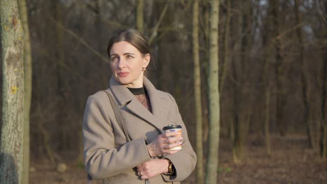 Middle-aged woman in light coat enjoys hot coffee in park.