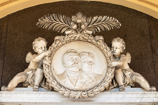 Round medallion with old couple held by baby angels carved in white marble. Close-up of tombstone at Vienna Central Cemetery old arcades with graves from 1880s