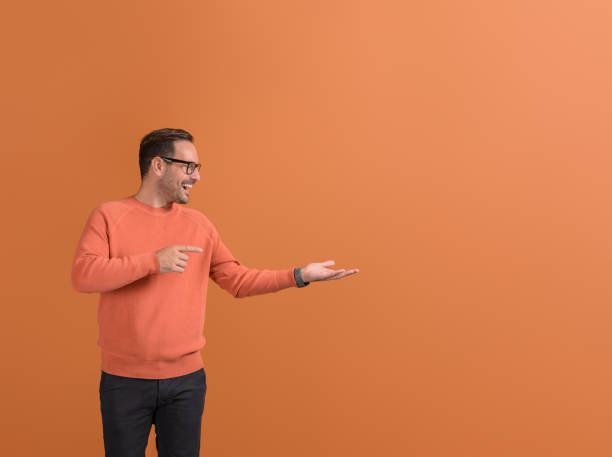 istock Ecstatic salesman pointing at empty palm and demonstrating new product on isolated orange background 2147585175
