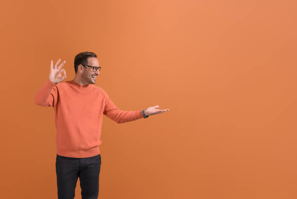 istock Happy young salesman showing OK sign and looking at empty palm while standing on orange background 2147585186