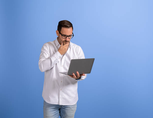 istock Serious young businessman with hand on chin thinking while working over laptop on blue background 2147585226