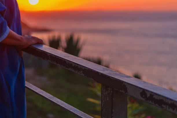 Handrail on which an elderly unrecognizable man leans with his hands stands against the background of an amazing sunset over the sea horizon, enjoying the magnificent landscape, view and life.