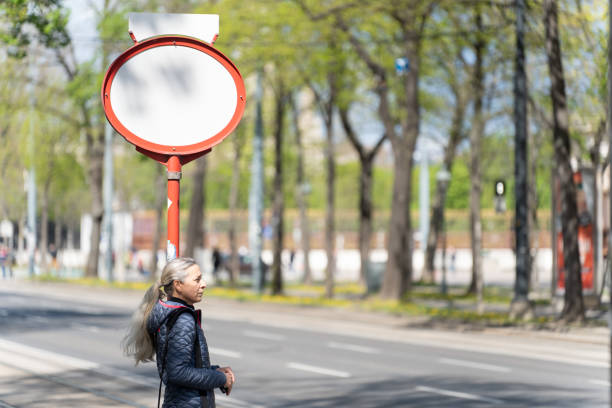 one mature adult woman waiting at tram stop in city of vienna on partly sunny spring day, background blurred, shallow focus on person, place for text on white sign
