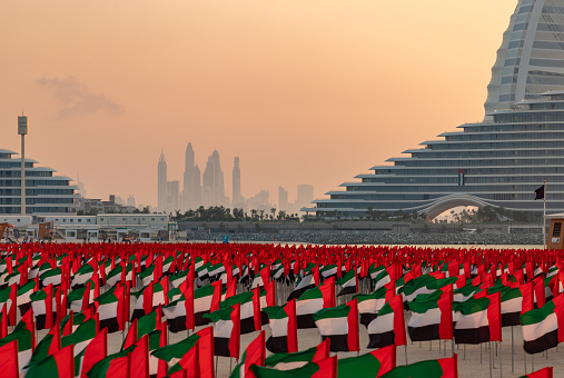 Dubai, United Arab Emirates - November 8, 2023: A picture of many United Arab Emirates flags at the Jumeirah Public Beach, with the Dubai Marina on the far left, at sunset.