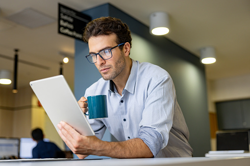 Latin American businessman at the office reading on a digital tablet using eyeglasses and drinking coffee