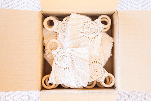 Elegant beige macramé napkin holders featuring a handcrafted pattern and wooden rings carefully packaged in a cardboard box for shipping showcasing their intricate design and craftsmanship. Part of a series.