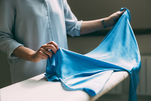 A mature woman carefully ironing and folding a bright blue piece of fabric, doing laundry chores at home.