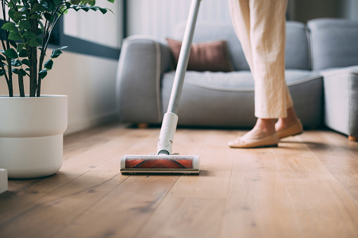 Close-up view of a person using a cordless vacuum cleaner on a wooden floor in a well-lit living room.