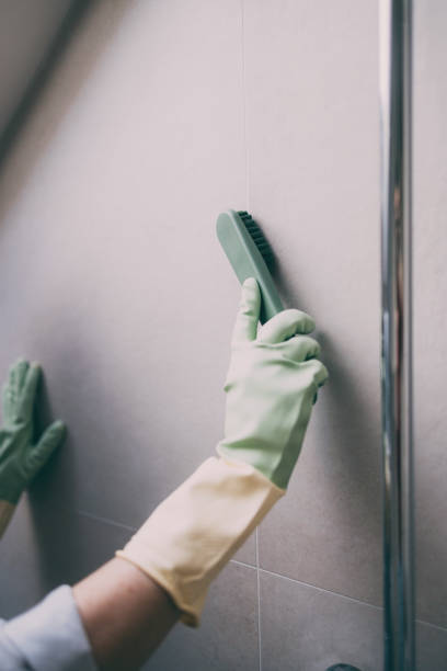 Close-up of hands in rubber gloves cleaning a tiled wall using a scrub brush. Concept of household chores and cleanliness.