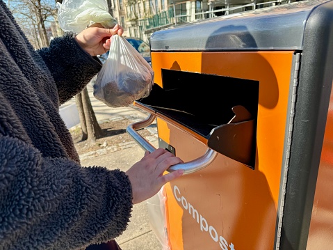 Close-up of young woman's hands and her shadow throwing a compostable plastic bag filled with food scraps into an orange public Smart Compost Bin with the word 