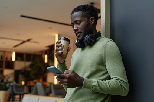 Minimal side view portrait of Black young man using smartphone leaning on wall in office lounge and drinking coffee copy space