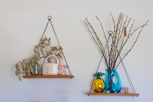 two triangular wall shelf with hanging vases and willow branches, one on the left is holding white tea flowers in ceramic mug, other has colorful blue glass vase with small flower ornament, wooden shelves, against plain light grey background, natural lighting.