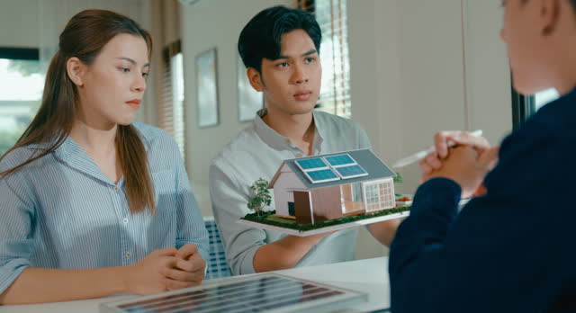 Closeup of young Asian couple discussion real estate agent about financial and environmental benefits of investing in home with solar technology. Eco-Friendly Home with Real Estate Agent.