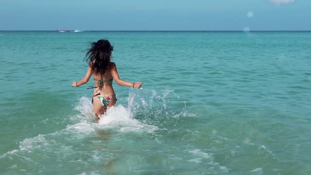 Slow motion of asian woman wearing beautiful colorful bikini run by beach at golden sunset - Female tourist on summer vacation. Pretty girl run to the sea splashing water waves. Crystal blue water.