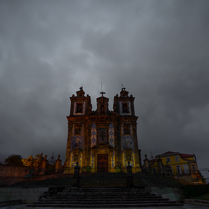 Church of San Ildefonso and steps lined with blue and white ceramic tiles wonderfully and dimly illuminated by orange lights at twilight, under a gray cloudy sky. Portugal.