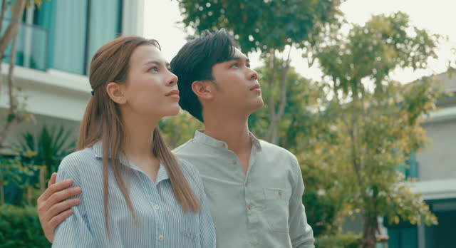 Closeup of Asian real estate agent demonstrates solar panel benefits to interested young couple outside a modern home. Consulting Eco-Friendly Home with Real Estate Agent.