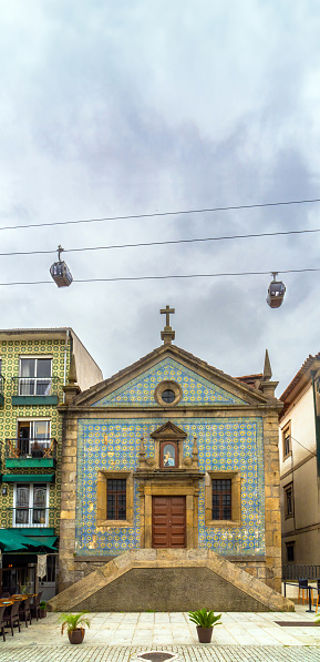 Chapel of Our Lady of Mercy in Vila Nova de Gaia, decorated with tiles, with two gondolas of the Gaia cable car passing over the Christian cross and roof. Porto Portugal.