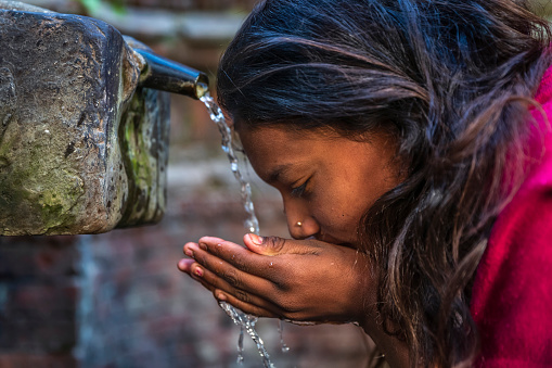 Young Nepali girl drinking from city fountain in Bhaktapur Nepal. Bhaktapur is an ancient town in the Kathmandu Valley and is listed as a World Heritage Site by UNESCO for its rich culture, temples, and wood, metal and stone artwork.