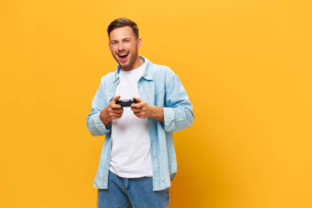 istock Cheerful young tanned handsome man in blue basic t-shirt play game hold joystick posing isolated on orange yellow studio background. Copy space Banner Mockup. People lifestyle Lucky winner concept 2147609130