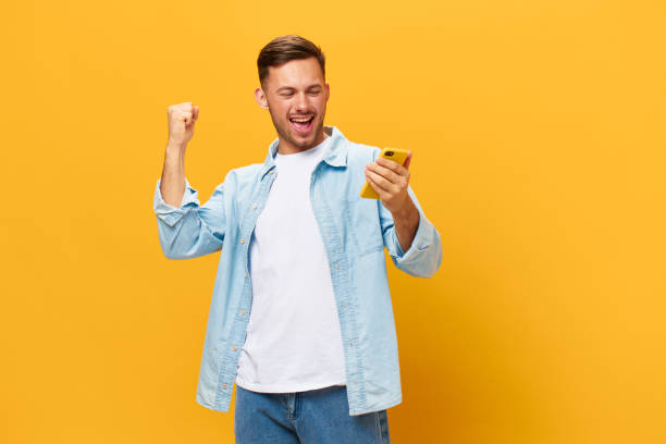 istock Smiling enjoyed tanned handsome man in blue basic t-shirt raise fist up using phone posing isolated on orange yellow studio background. Copy space Banner Mockup. Online Social media. Lucky win concept 2147609528