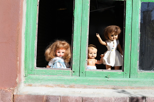 A spooky set of ancient dolls are looking through a window that seems to belong to a secret and abandoned place in the middle of nowhere. The setting is perfect for a Halloween decoration