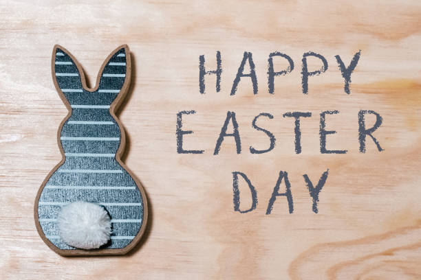 A rabbit is on a wooden surface with the words Happy Easter Day written below it. The rabbit is decorated with a white pom pom on its back