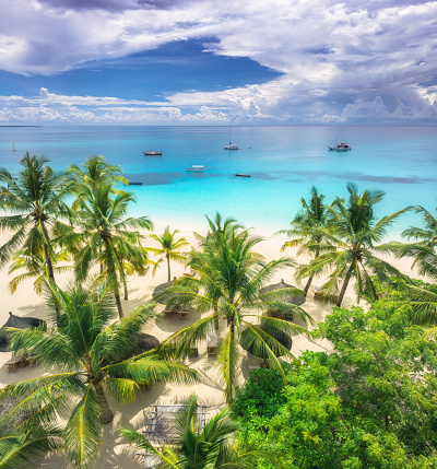 Aerial view of white sandy beach with palm trees, umbrellas, yachts, boats, blue ocean, sky with clouds at sunset. Summer vacation in Kendwa, Zanzibar island. Tropical landscape. Clear sea. Top view