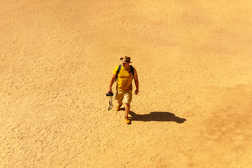 Man walking in the middle of desert in Egypt, Africa. He is wearing a hat, yellow t-shirt and shorts. He holds a professional camera in his hand
