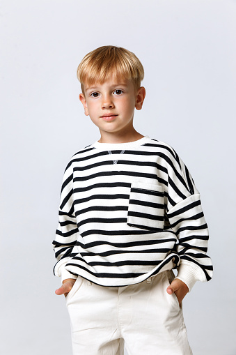 Blonde little kid boy model in striped sweater and white trousers posing on white studio background. Fashion kid model
