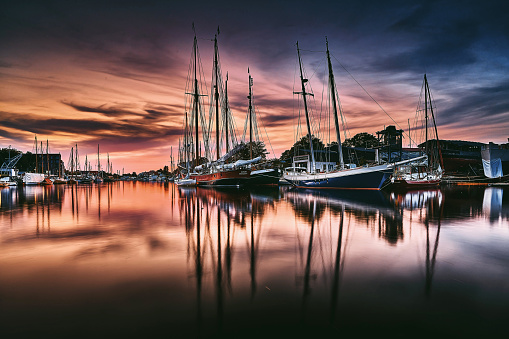 Picturesque scene where ships rest in the harbor, and the evening light bathes the sky in warm tones.

At the center of attention are the moored sailboats, gently rocking on the water. The masts stand sharply against the overcast sky, and the reflections of the ships on the calm water surface add an extra layer of beauty.

The evening light transforms the scene into warm oranges and soft blue and gray hues. The sky is adorned with clouds that capture the light of the setting sun, creating a special atmosphere of tranquility and contemplation.

The harbor surroundings exude a serene calmness, and the moored sailboats appear as guardians of the maritime ambiance. The scene radiates a timeless elegance typical of harbor evenings when the world settles into peace, and the maritime magic reaches its peak.

This artwork is ideal for creating a relaxed and inspiring atmosphere in hotels, waiting rooms, hallways, or vacation homes. It becomes a timeless reminder of the beauty of harbor evenings and the unique mood they evoke.