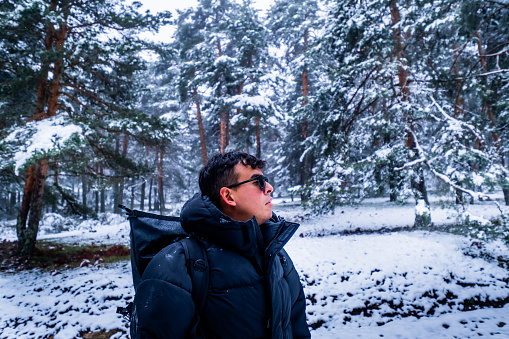 Wide-angle shot captures Latino Peruvian youth in black sunglasses and coat, admiring snowy forest.
