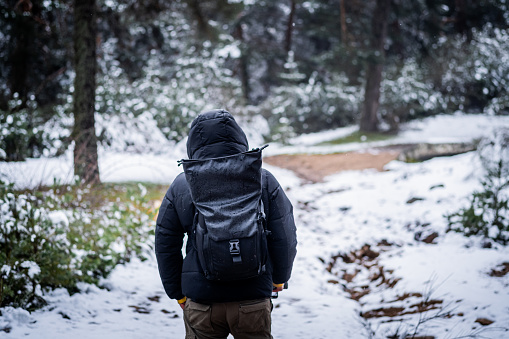 Back view photo of unrecognizable young man in black coat, hood, and backpack hiking snowy trail.