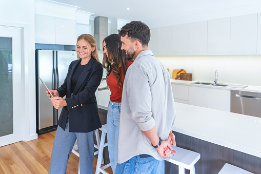 Real estate agent showing a couple a new home. The agent is holding a digital tablet which everyone is looking at. The kitchen can be seen in the background.