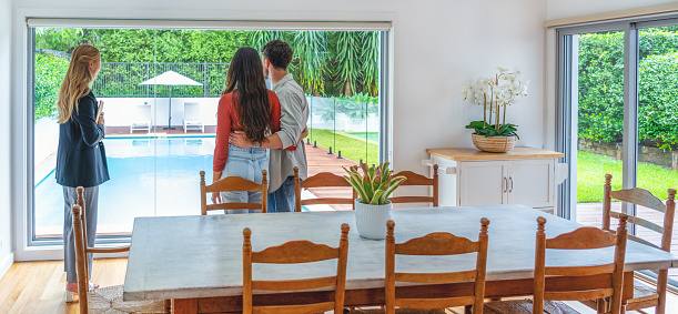 Real Estate agent showing a couple through a new house. They are standing in the dining room. She is showing the swimming pool and outdoor area through a window
