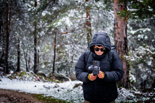 Snow-covered forest, tall pines, a Latino youth in black, filming with gimbal, showing love nature