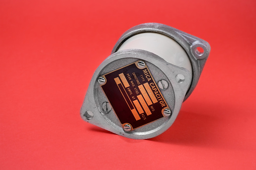 antique high voltage mica capacitor with a metallic casing and label detailing its specifications; it’s placed against red background