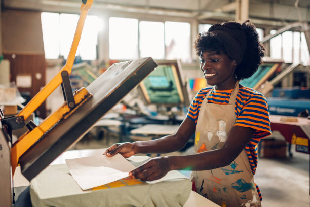 Smiling interracial printing shop worker preparing product for heat press Happy multicultural printer woman preparing t-shirt for heating press. Smiling african american printing workshop female worker standing at facility and putting paper on silk screen printed product. t shirt transfer stock pictures, royalty-free photos & images