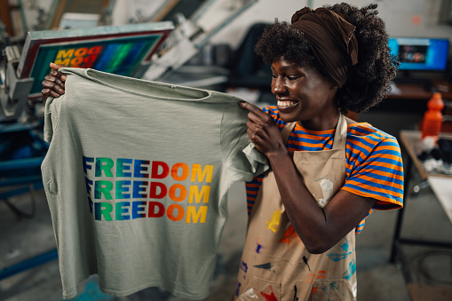 Multicultural printing shop female worker standing at facility with silkscreen printed finished textile product and showing it at the camera. Portrait of a smiling interracial press woman with t-shirt